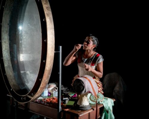 Deaf Role Model Nadia on stage, looking at a mirror in front of a box with a number of items