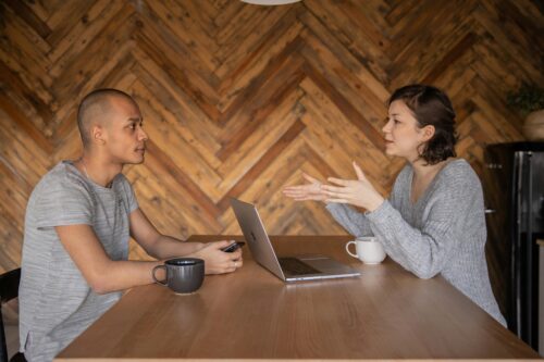 Two people sit opposite each other at a table, with a laptop and coffee cup, in a meeting.