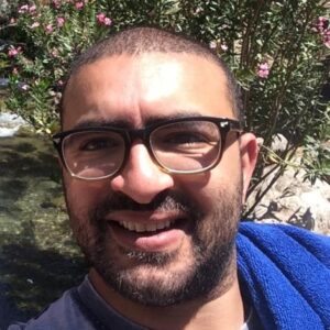 Bassim Baz smiling with short hair, a beard, and glasses, standing outdoors in front of a bush with pink flowers.