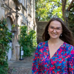 Kirstie is smiling at the camera. She is stood outside, with an old stone college building in the background with some plants growing and a lamp directly behind her. Her brown hair is flowing in the wind. She has some brown glasses, moonstone earrings, and a vibrant patterned V-line dress with shades of red, pink and blue.