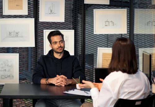 A South Asian man sits at a desk opposite a woman with shoulder length black hair, in an interview setting