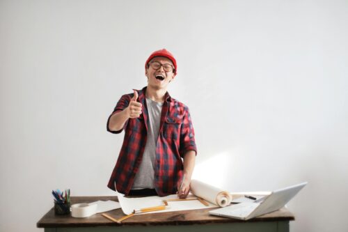 A man in a hard hat with a checked shirt stands at a desk with pencils, paper and measuring material. He has one hand in a thumbs up gesture and is smiling.