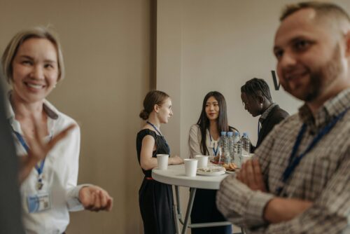 In the foreground a man and woman wearing lanyards smile welcomingly at someone offscreen. In the distance, 3 more people wearing business attired and lanyards have a discussion.