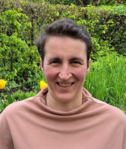 A woman with short hair smiles at the camera in front of greenery