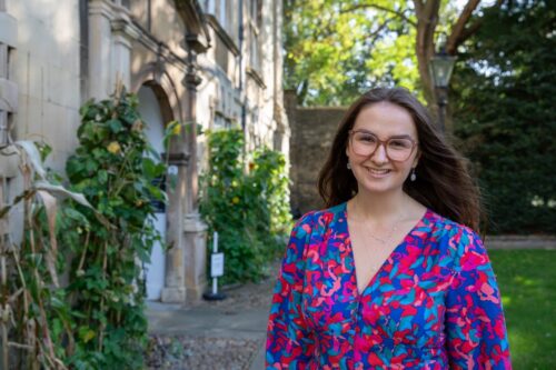 An image of Kirstie smiling at the camera, with her Cambridge college in the background. She is wearing a multicoloured V-neck dress, moonstone earrings, glasses. Her brown hair is flowing gently in the wind and there are some plants and trees behind her.