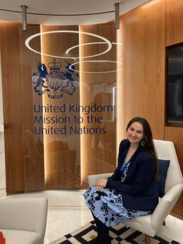 Kirstie is wearing a navy blazer and a light-blue and black dress with light blue earrings. She is sat on a cream leather seat with the glass panel that reads ‘United Kingdom Mission to the United Nations’ and has the UK crest.