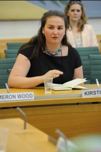 A very young Kirstie is talking in a parliamentary select committee room in the Houses of Parliament. She has a notebook in front of her and her name tag. There is a person behind her. She is sat down, wearing a formal black dress, a circular blue and purple necklace, silver hoop earrings and pulled back hair.